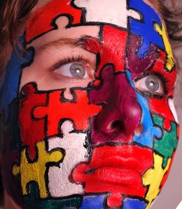 woman's face with red, yellow, white, green and blue puzzle pieces painted on, with a missing piece around her right eye