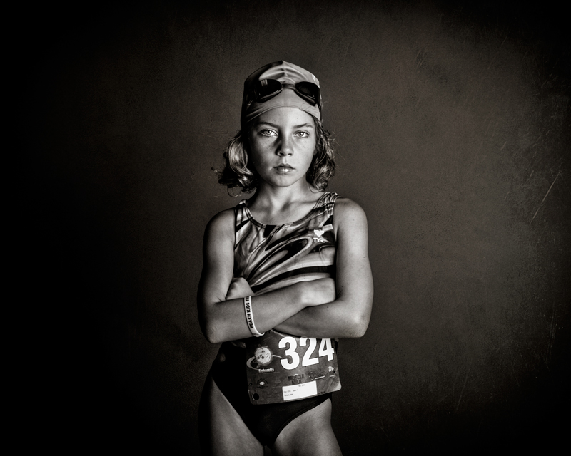 black and white photo of a preteen female athlete in a swim suit, swim cap, goggles and a race number pinned to her suit. 
