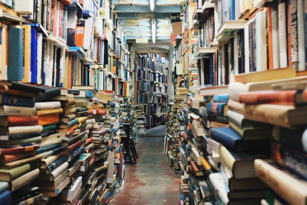 a narrow hallway filled to the brim with books, from floor to ceiling. Probably is a basement in a library. The books are piled haphazardly.